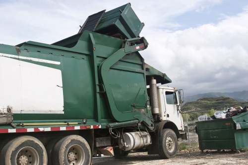 A large green waste collection truck with a hydraulic lift mechanism is parked on a gravel surface, partially lifting a green skip at an angle with its open top revealing waste materials inside. The truck's cab is white with a visible side mirror and black tires, positioned on the right side of the image, facing towards the background which features a scenic landscape of rolling hills and a cloudy sky. To the right of the truck, there is a green waste bin filled with mixed rubbish, including plastic bags and assorted debris, situated outdoors in an open area near Hampstead. The scene suggests a waste removal operation in a rural or semi-rural setting, typical of local rubbish collection services offered by Business Waste Removal Hampstead, serving the Hampstead postcode area.