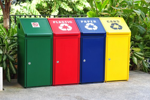A row of four large, upright waste recycling bins positioned on a paved outdoor surface, with lush green foliage and plants in the background. The first bin on the left is green with a slot for general waste, featuring a small symbol indicating no food waste. Next to it, the red bin is designated for plastic recycling, with a white recycling symbol and the word 'PLASTIC' printed above. The third bin is blue, intended for paper recycling, marked with a white recycling symbol and the word 'PAPER.' The fourth bin on the right is yellow, designated for cans, with a matching recycling symbol and the word 'CANS.' Each bin has a simple door with a small handle at the front and a circular lock mechanism. The overall scene suggests a tidy, organized waste separation point likely situated outside a premises in Hampstead, supporting effective rubbish management services offered by Business Waste Removal Hampstead. The setting is well-lit, with natural daylight illuminating the vibrant colors of the bins against the natural greenery, contributing to environmentally conscious waste collection in the local area of NW3 postcode.