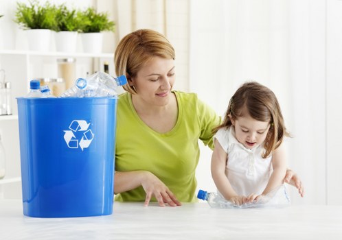 A woman with short, light brown hair, wearing a bright yellow-green top, is engaging with a young girl with shoulder-length brown hair dressed in a white top. They are indoors in a well-lit, modern space with a white background featuring open shelving filled with white pots containing green plants. On a smooth, light-colored surface in front of them, there is a large blue recycling bin with a white recycling symbol, filled with crumpled, clear plastic bottles of various shapes and sizes, some with blue caps. The woman appears to be guiding the girl in sorting or examining the plastic waste. The scene emphasizes waste management and recycling, reflective of a professional rubbish removal service in Hampstead, with the environment clean and organized, highlighting an awareness of proper waste disposal practices.